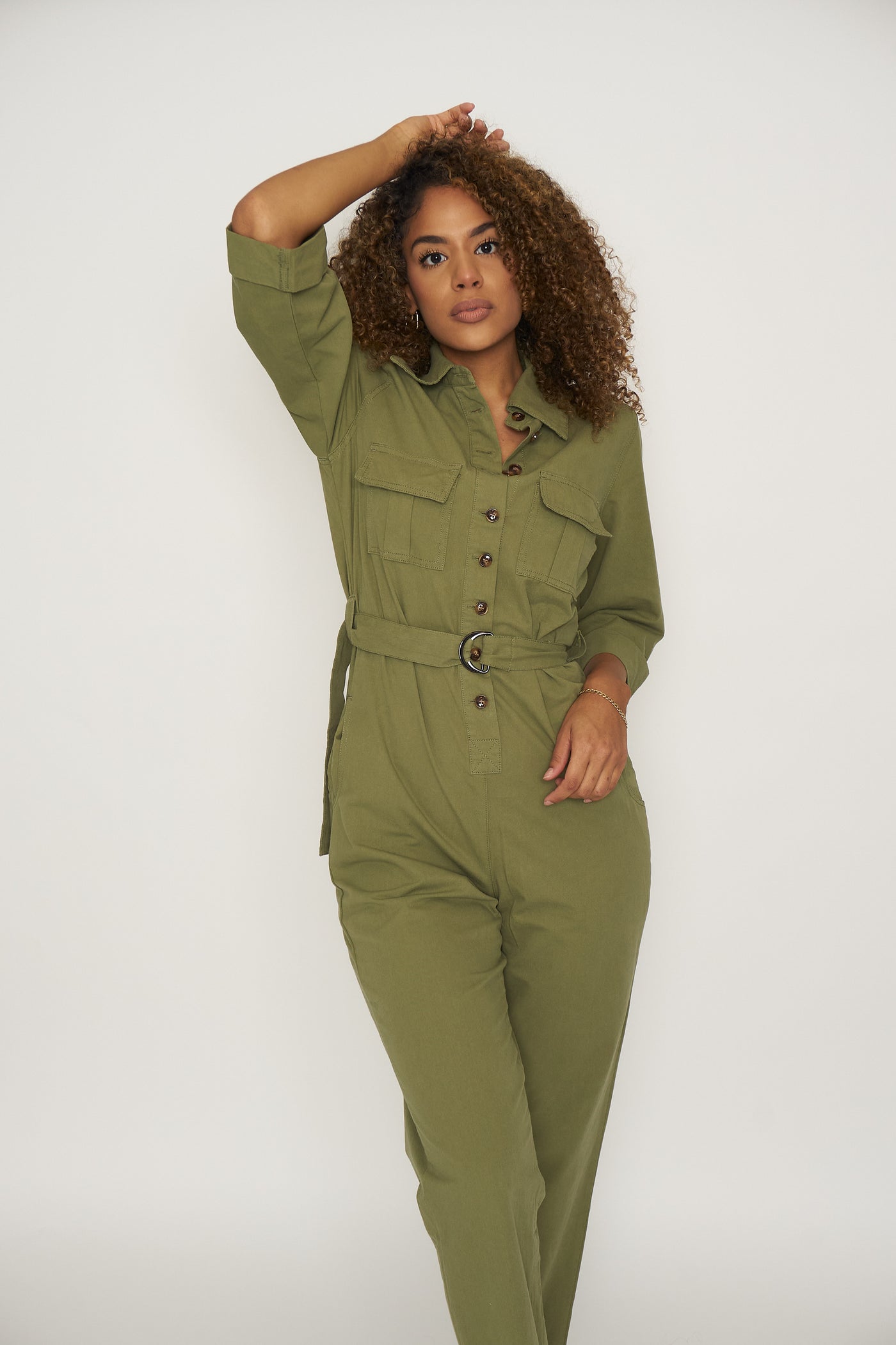 Woman wearing an olive green utility jumpsuit. The jumpsuit has brown tortoise button down the front until the waistline, 2 breast and 2 side pockets, rolled quarter sleeves, collared neckline and D-king belt 