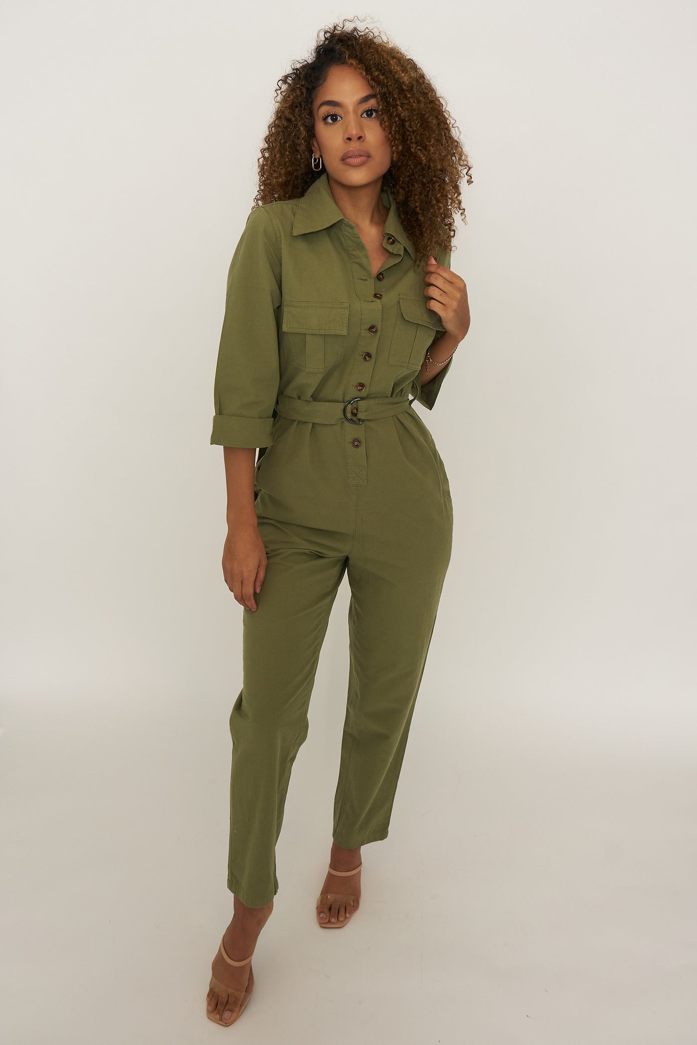 Woman wearing an olive green utility jumpsuit. The jumpsuit has brown tortoise button down the front until the waistline, 2 breast and 2 side pockets, rolled quarter sleeves, collared neckline and D-king belt 