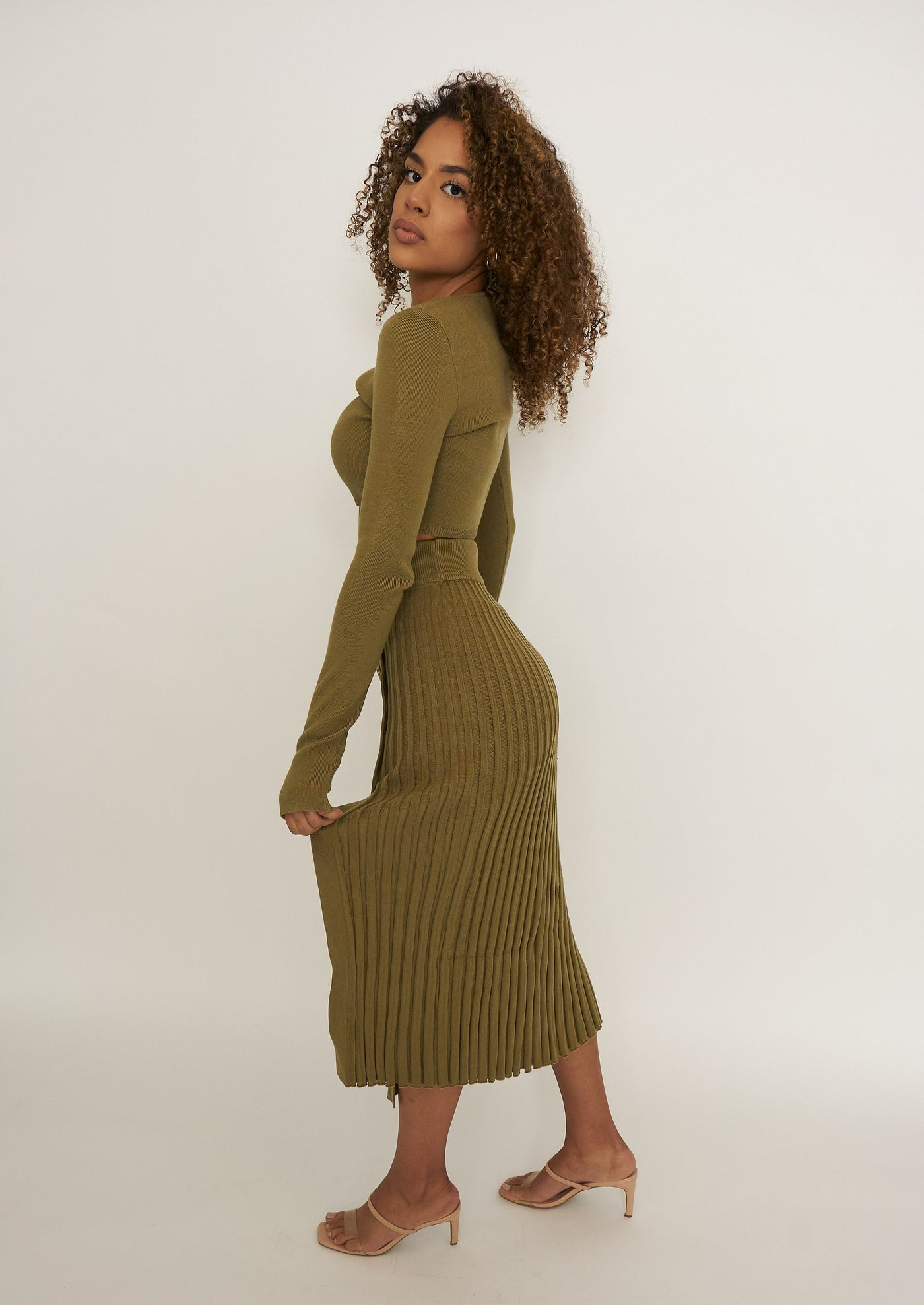 Show Me Leg is an olive green sultry pleated, mid-thigh slit sweater skirt paired with a long sleeve crop top. The crop top has a thumb hole feature that allows for easy layering and provides a sleek look. The skirt has a natural hourglass shape that helps accentuate your hips, slims your waist creating the hourglass effect when put on.